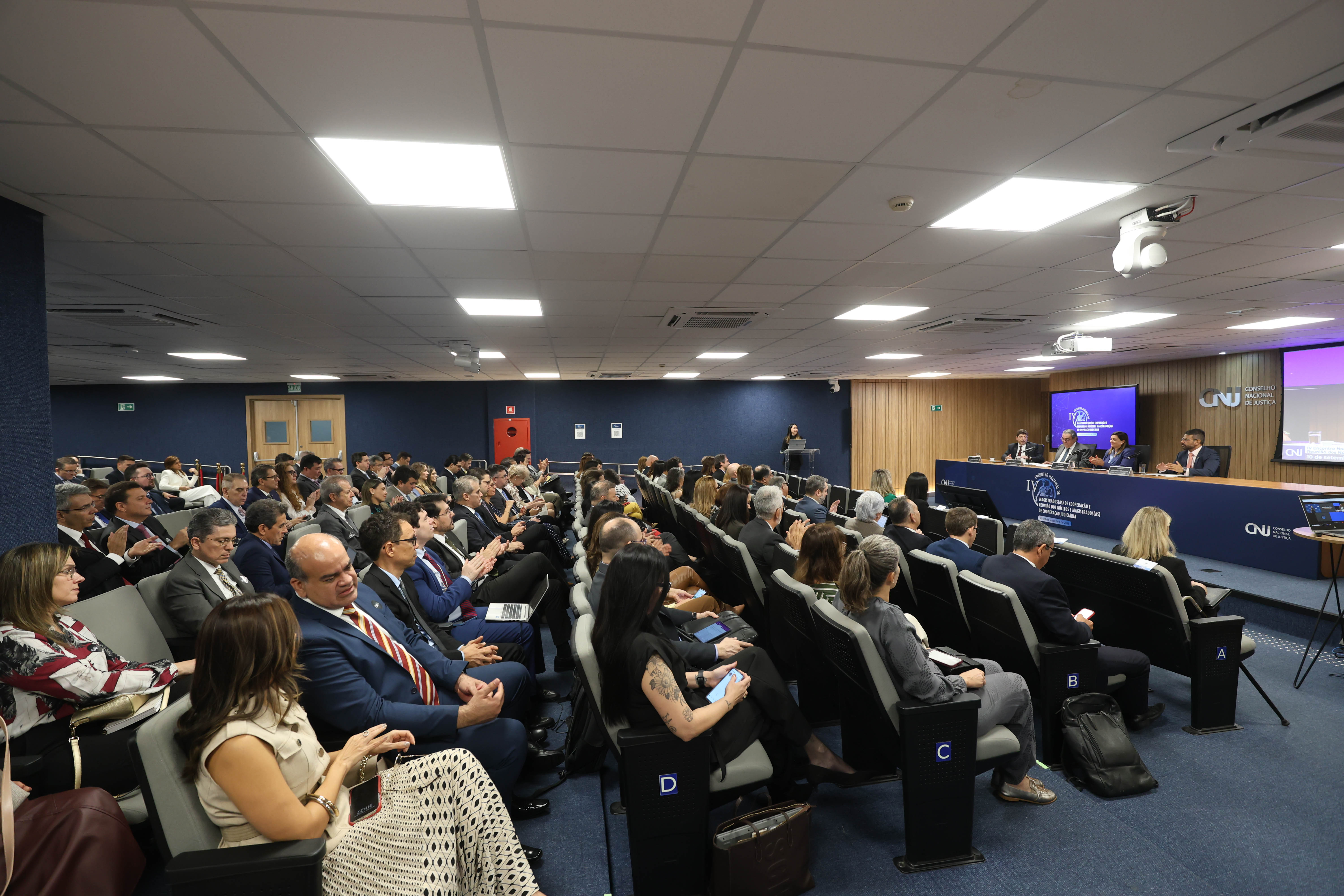 Imagem de uma sala de conferência com pessoas assistindo a uma sessão do CNJ, com painel, telão e algumas usando celulares.