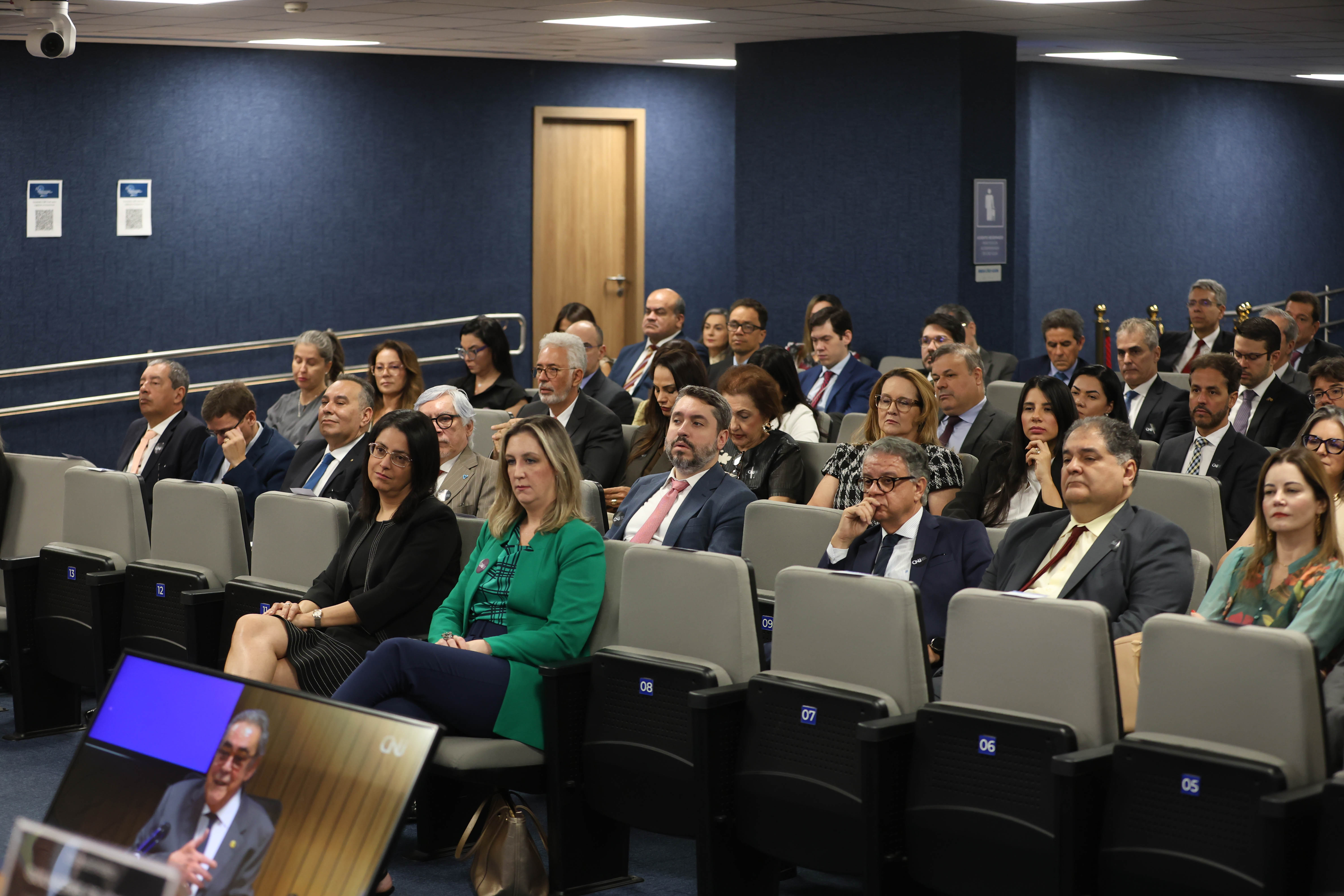 Reunião formal com várias pessoas sentadas em auditório, assistindo a uma apresentação em tela, entre elas a juíza Adriana