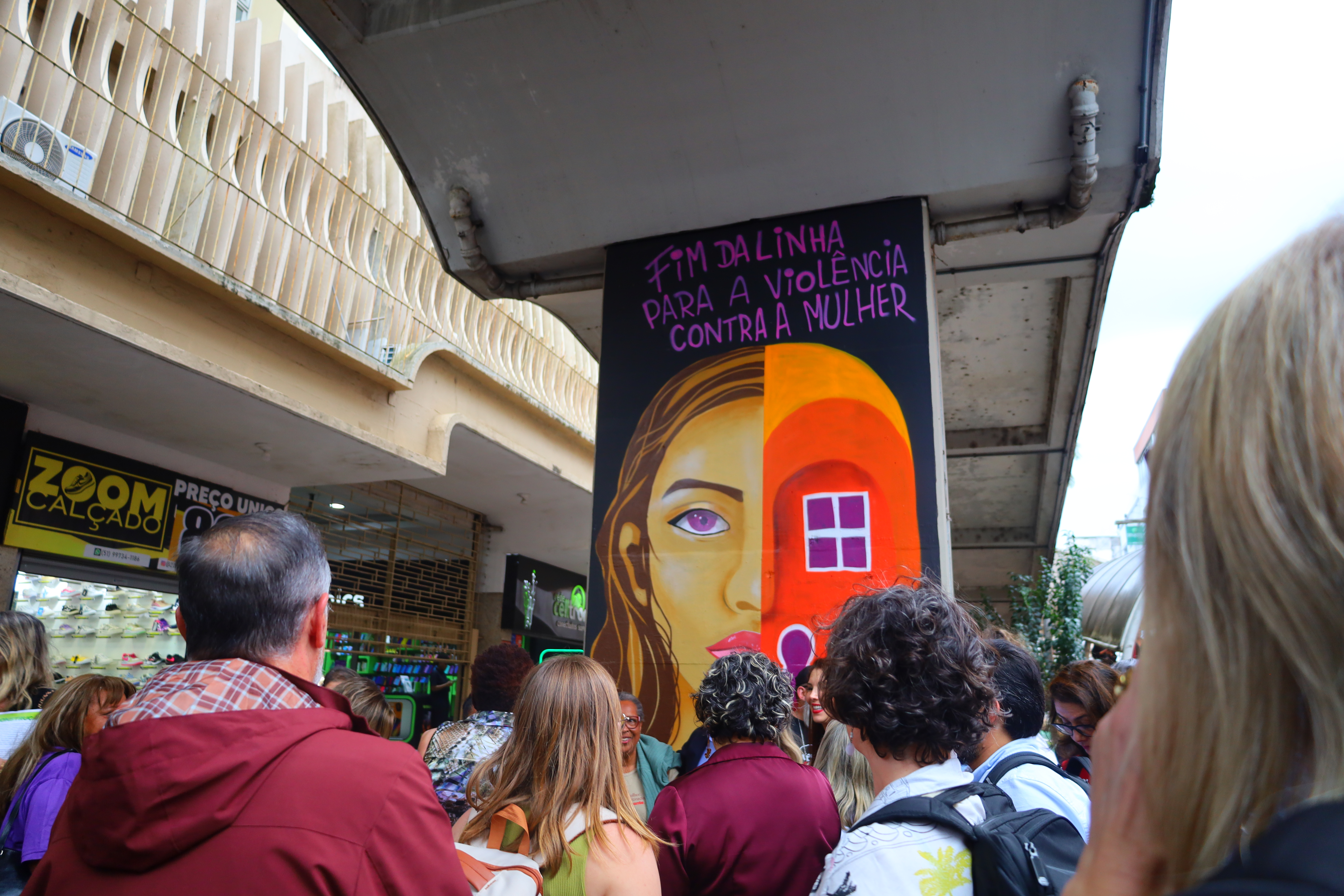 Arte em grafite na saída da Estação Canoas, com o rosto de uma mulher e a frase “Fim da linha para a violência contra a mulher”. Um grupo de pessoas observa a obra.