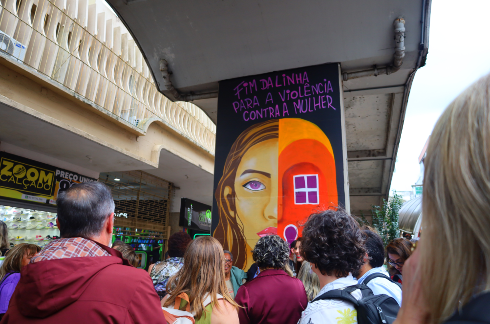Arte em grafite na saída da Estação Canoas, com o rosto de uma mulher e a frase “Fim da linha para a violência contra a mulher”. Um grupo de pessoas observa a obra.