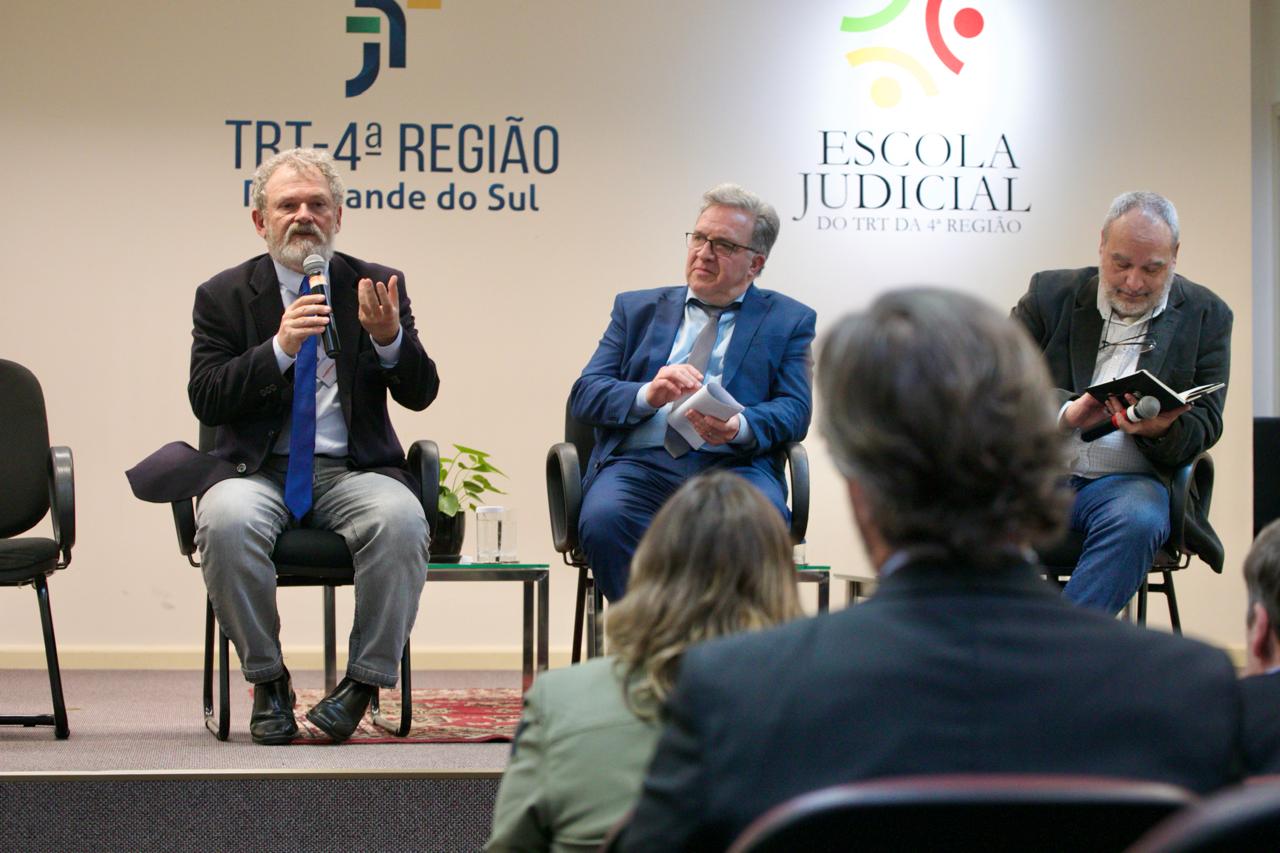 A foto mostra três pessoas no palco, um delas falando ao microfone.