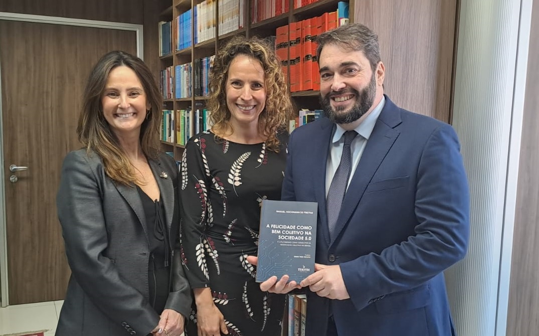 A foto mostra três pessoas posando para foto com um livro em mãos.