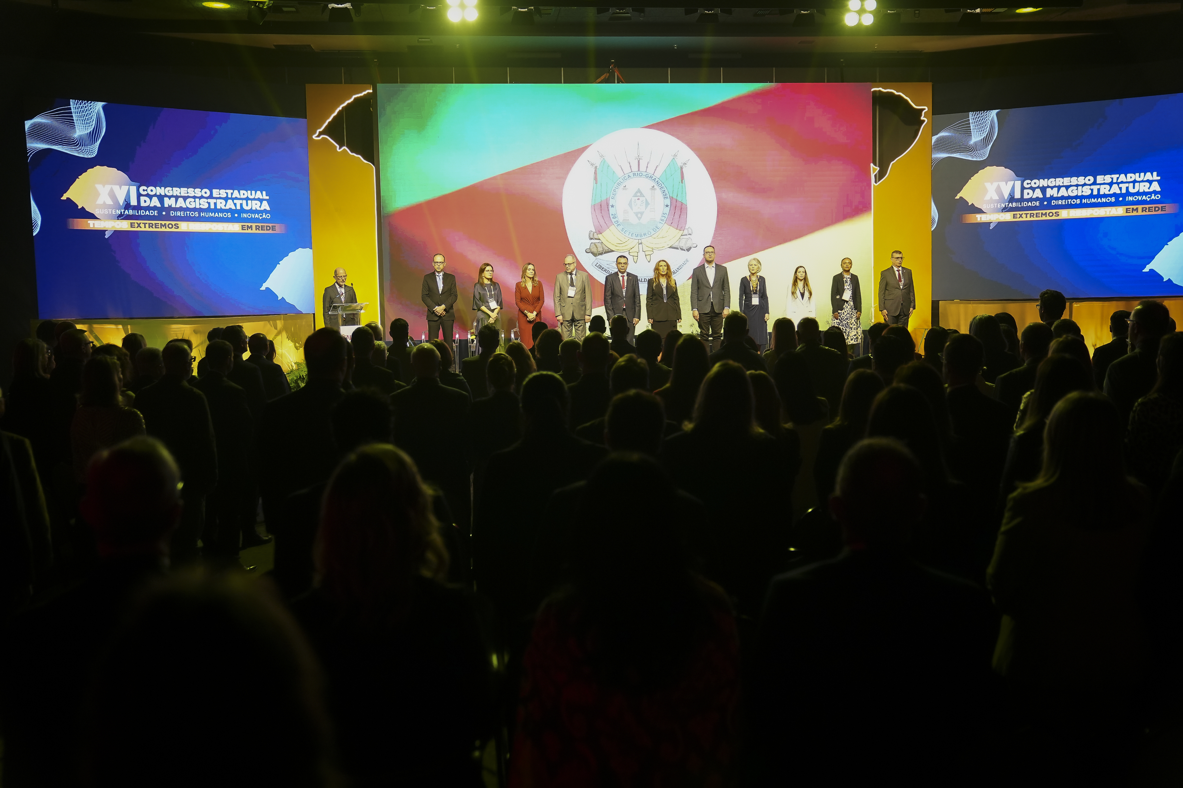 Mesa de abertura do Congresso da AJURIS. Os integrantes estão em pé, atrás deles há um telão exibindo a bandeira do RS.