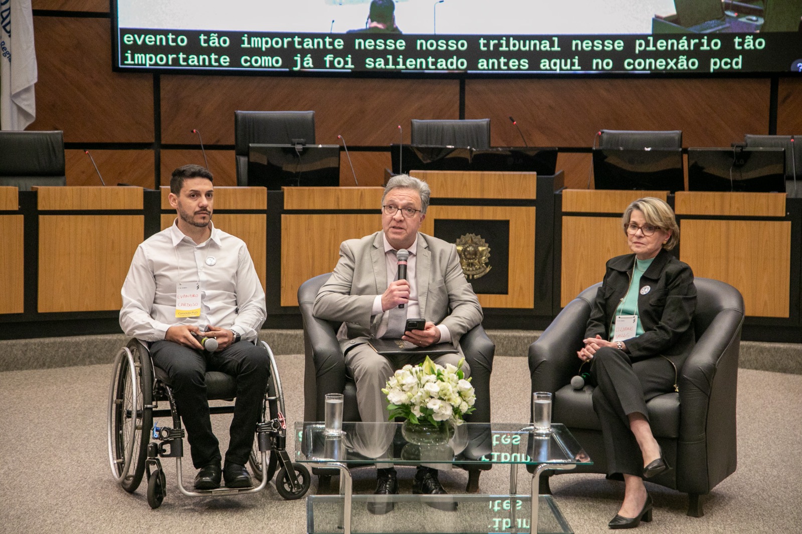 Três palestrantes (do painel 2) sentados em poltronas: um homem em cadeira de rodas (Evandro), outro de terno cinza falando ao microfone (Cassou) e uma mulher de óculos e casaco preto (Liliane). À frente, mesa de vidro com flores e copos de água.