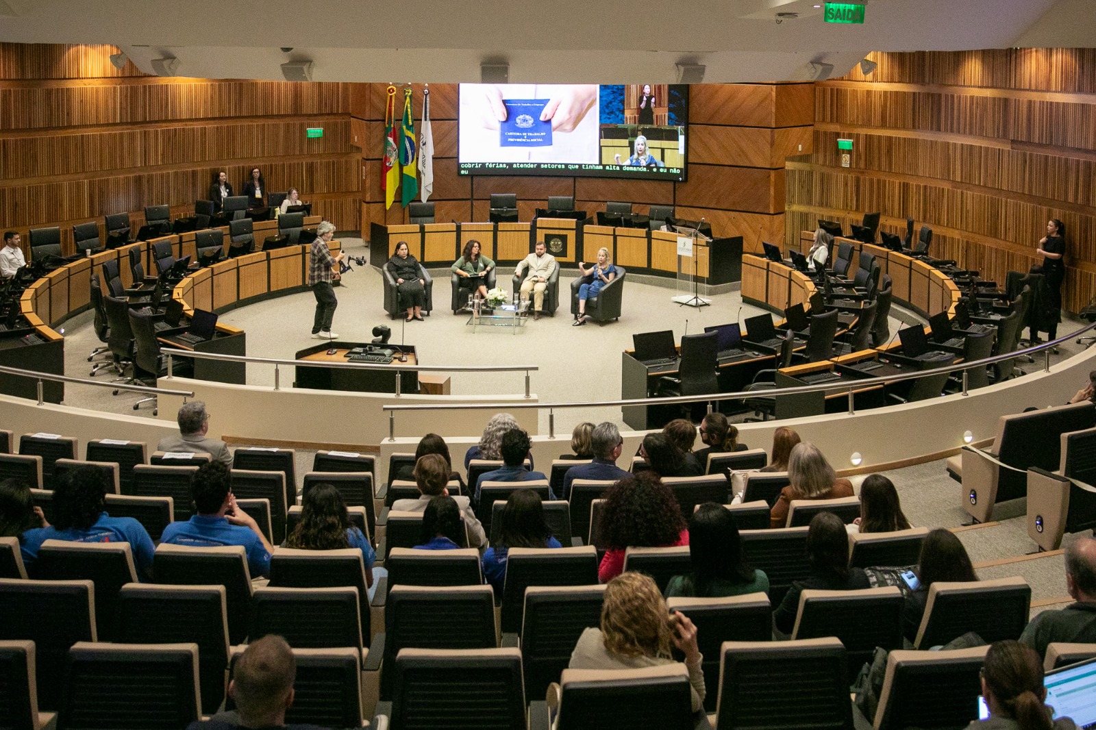 Visão geral do plenário com público atento e painelistas no centro do espaço. O grande telão ao fundo exibe imagens do evento ao vivo.
