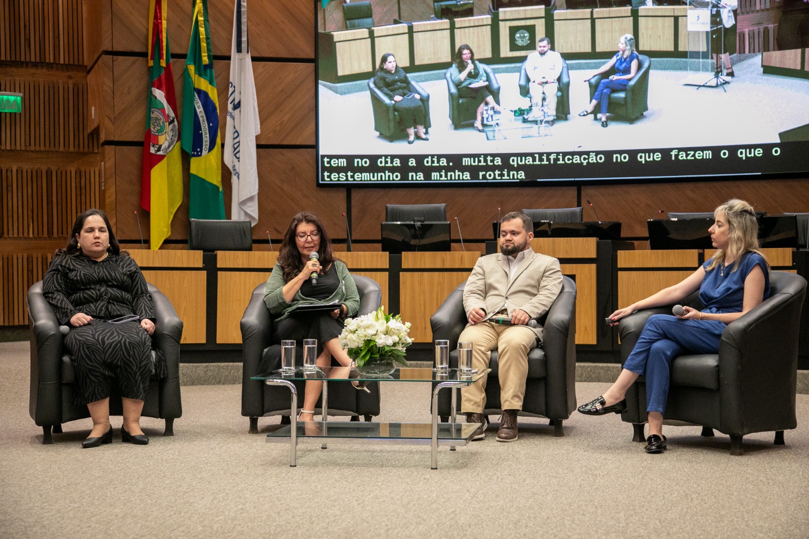 Quatro palestrantes estão sentados em poltronas pretas (do painel 1), dispostos em semicírculo. À esquerda, Juliana está de vestido preto com estampa animal print observa atenta. Ao lado dela, Luciana, de óculos e casaquinho verde, segura o microfone e fala ao público. À direita, Juliano, que veste um terno bege e sapatos marrons, escuta atento. Na ponta, Renata, que está de macacão azul, segura um microfone no colo, acompanhando a fala. À frente, mesa de vidro com flores brancas e copos de água