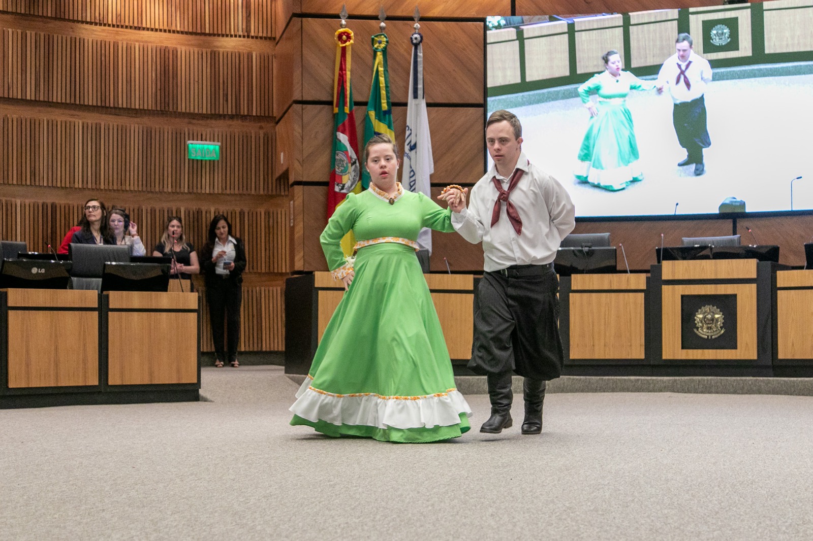 Casal de jovens em trajes típicos gaúchos dança no centro do plenário. A moça usa vestido verde longo e o rapaz, bombacha e lenço vermelho. Ao fundo, bandeiras e um telão exibem a apresentação ao vivo.