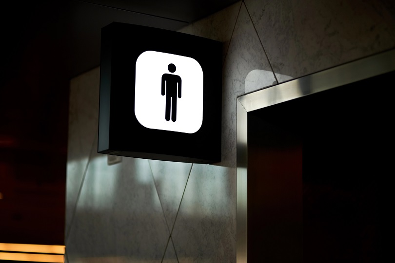A foto ilustrativa mostra a entrada de um banheiro masculino. Há uma placa iluminada com fundo branco e moldura preta, exibindo o ícone de uma figura masculina estilizada em preto.