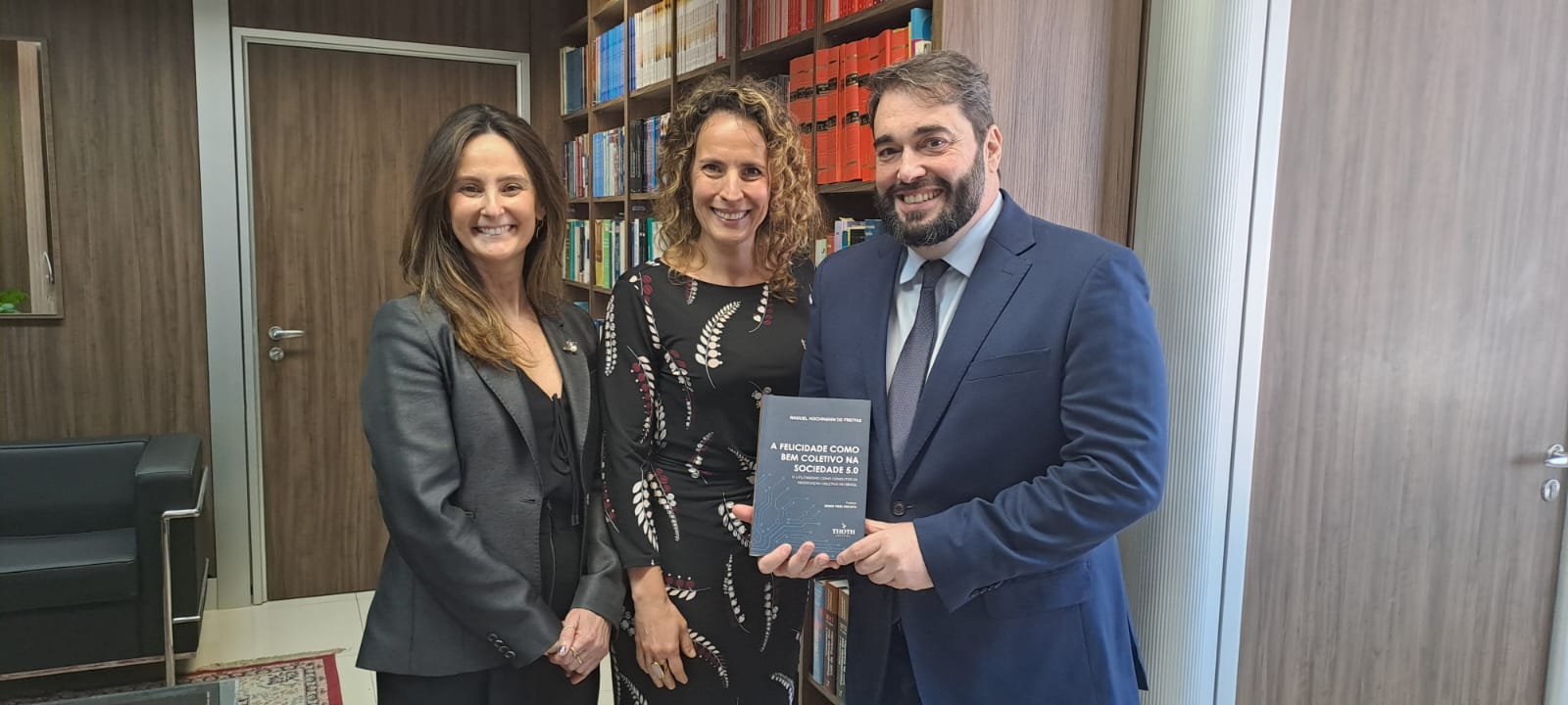 A foto mostra três pessoas posando para foto com um livro em mãos.
