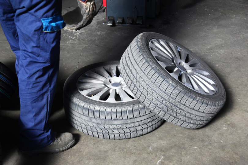 homem de uniforme azul está ao lado de um carro e dois pneus, que estão no chão em uma oficina. Não há imagem do rosto.