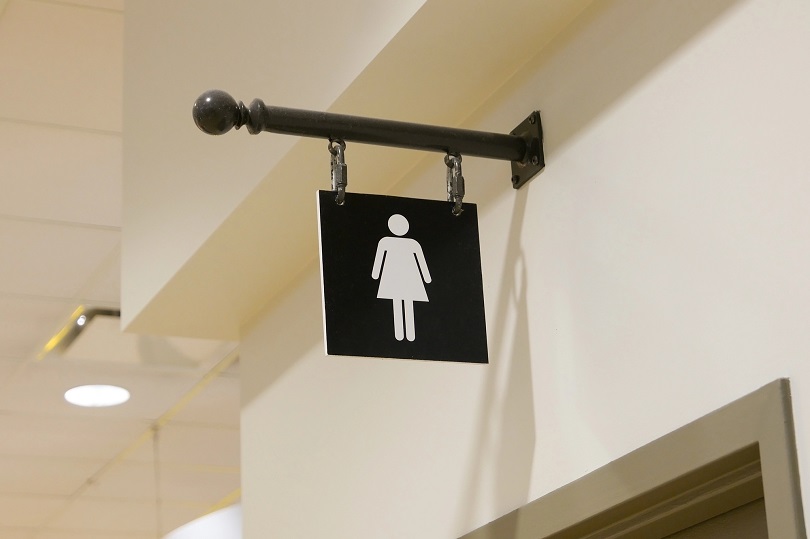 A foto ilustrativa mostra uma placa de banheiro feminino. Ela está presa a uma haste metálica preta, fixada na parede com suportes. A placa é quadrada, de cor preta, com o desenho em branco de uma figura feminina estilizada, usando saia, que é o símbolo universal para indicar banheiro feminino.