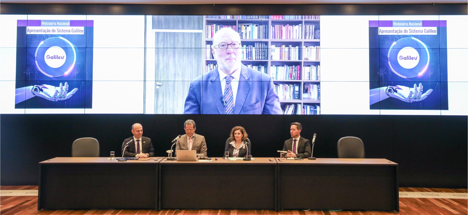 A foto mostra uma mesa de autoridades e o presidente martins costa de fundo, num telão.