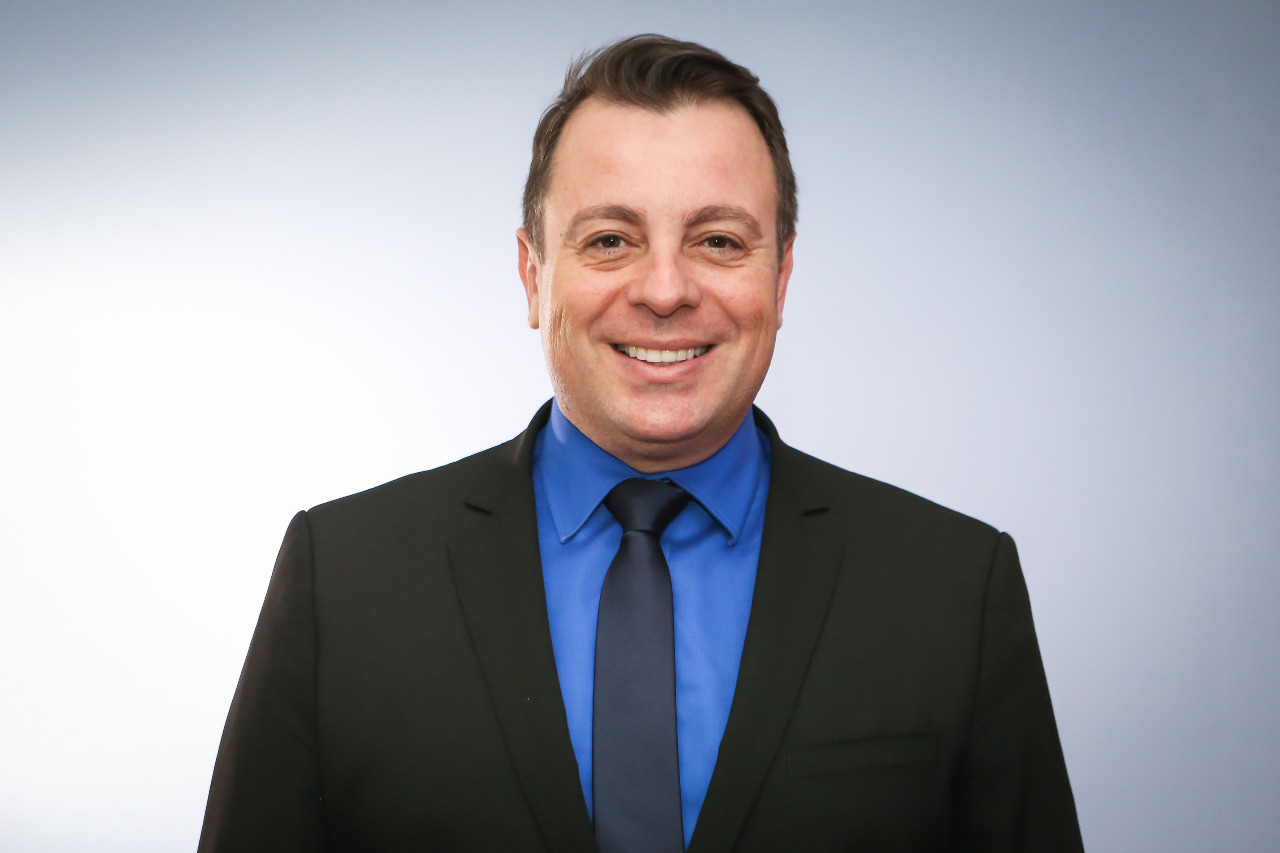Foto do juiz Márcio Lima do Amaral. Ele é branco, usa terno escuro, gravata escura e camisa social azul. Está sorrindo para a foto.