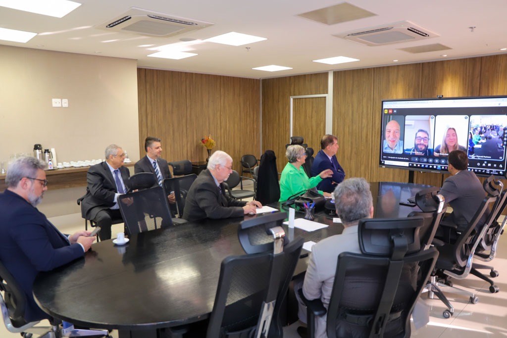 A foto mostra a mesa com pessoas em volta e um telão com outros participantes virtualmente.