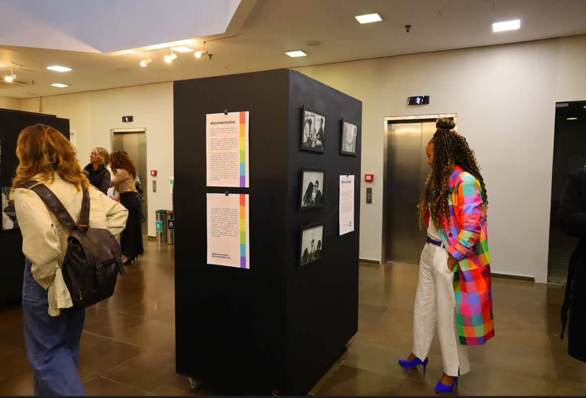 Pessoas circulando, olhando as fotos em painéis pretos. Uma mulher à direita veste casaco colorido quadriculado, calça branca e sapatos azuis, está observando a exposição.