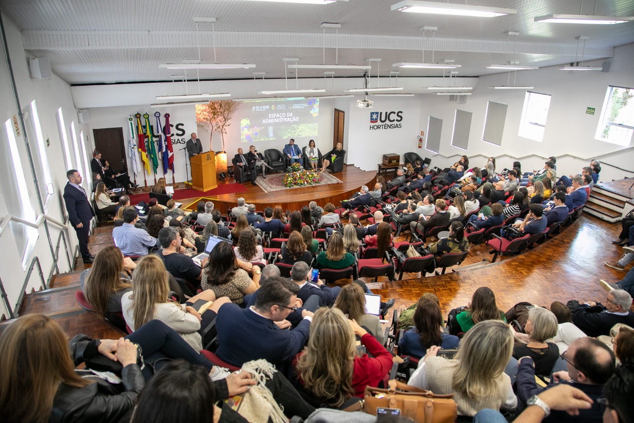 A foto mostra o público em primeiro plano e o palco com autoridades de fundo.