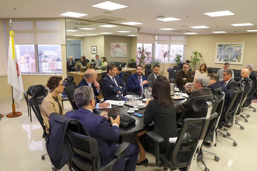 A foto mostra uma grande mesa e pessoas em volta dela numa reunião.
