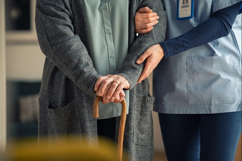 Cuidadora segura braço de idosa. Elas têm a pele clara e os rostos não são mostrados. A cuidadora veste uniforme azul e a idosa, roupa cinza