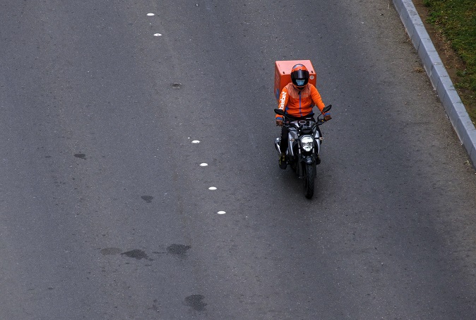 A foto ilustrativa mostra um motociclista trafegando sozinho por uma via asfaltada, visto de cima. Ele veste um uniforme laranja chamativo e capacete, indicando que provavelmente trabalha com entregas.