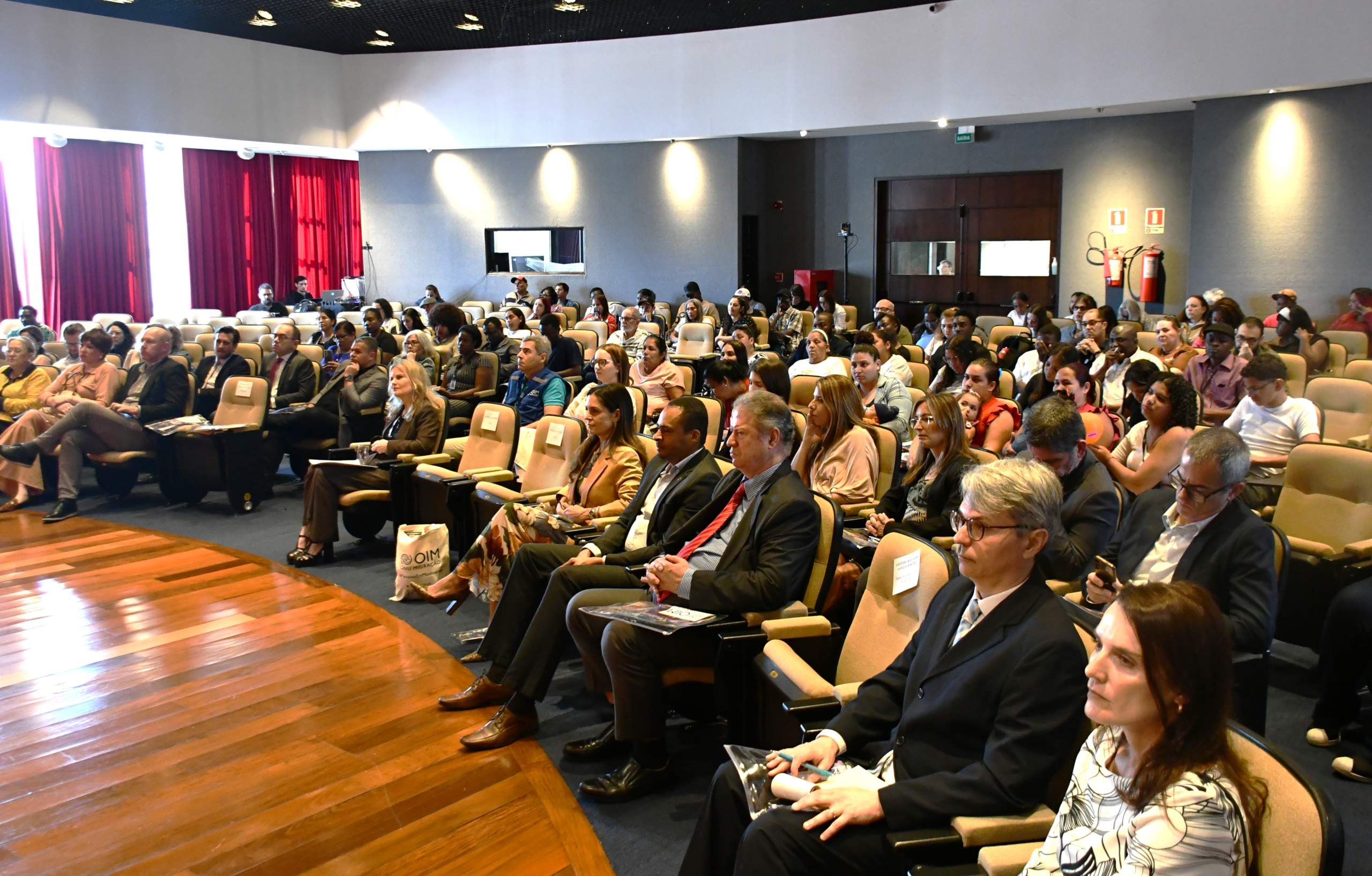 Auditório com público sentado em poltronas acolchoadas, voltado para um palco de madeira. A imagem mostra dezenas de pessoas acompanhando um evento, distribuídas em fileiras organizadas. Ao fundo, paredes claras com portas de madeira e extintores visíveis próximos à saída. Cortinas vermelhas cobrem as janelas à esquerda, permitindo iluminação natural combinada com luz artificial. O ambiente é amplo e bem iluminado, com disposição típica de espaço para palestras ou seminários institucionais.