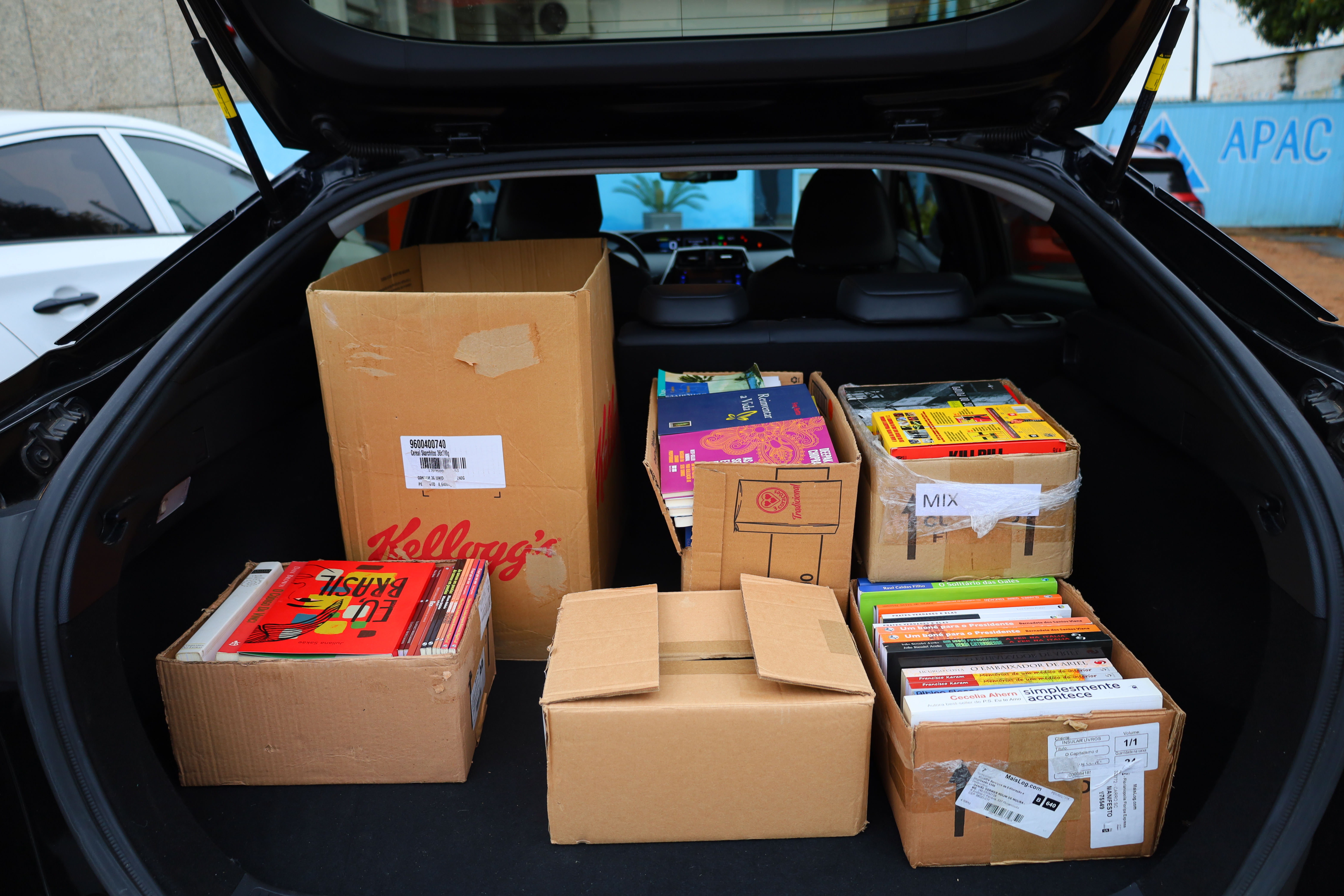 Porta-malas de um carro preto aberto com seis caixas de papelão, cheias de livros. Os livros estão organizados vertical e horizontalmente, com lombadas coloridas visíveis. O carro está estacionado em frente a um muro azul com a sigla "APAC".