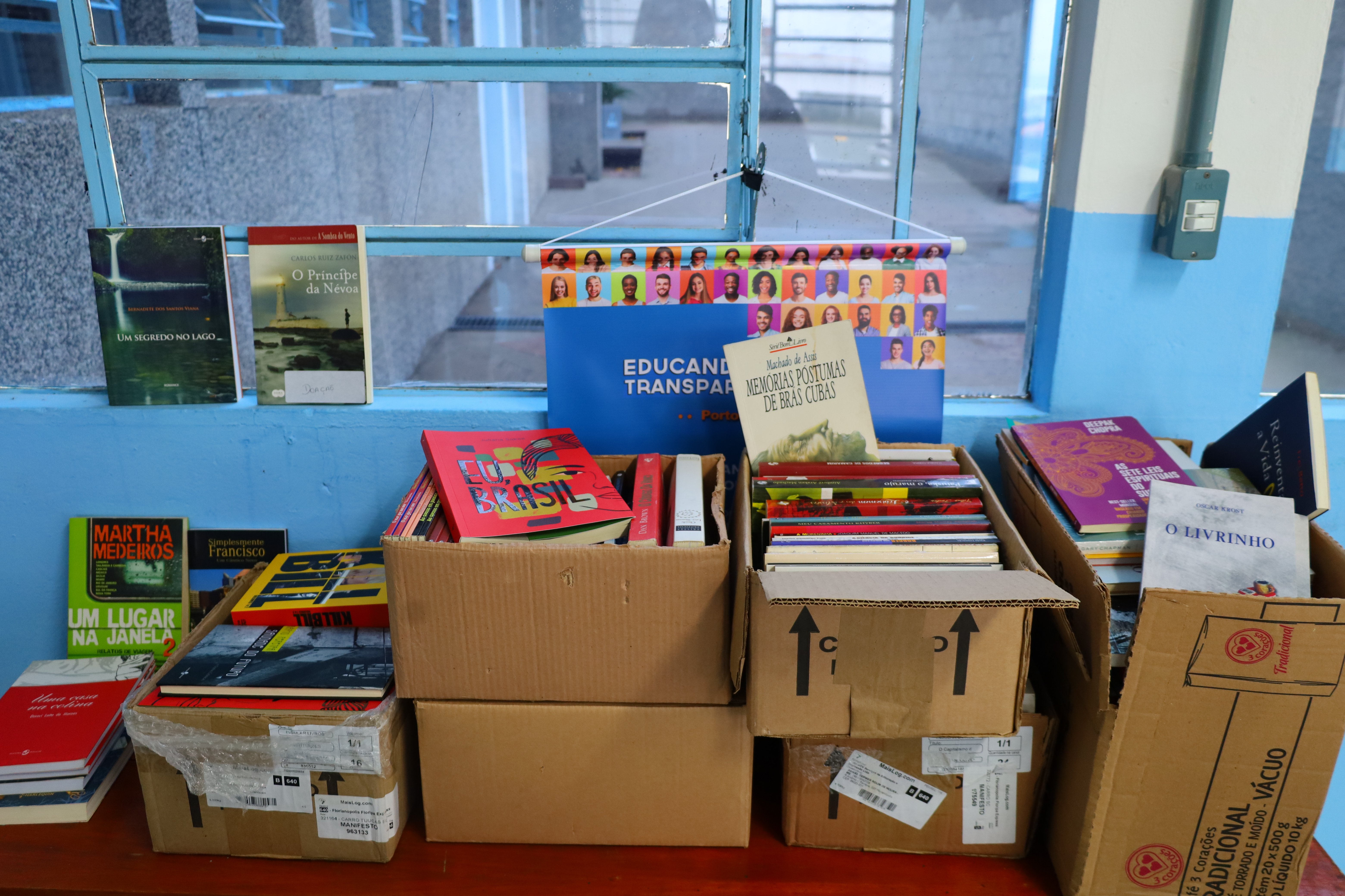 Caixas de papelão sobre uma mesa de madeira, todas abertas e com alguns livros arrumados em pé e empilhados. Ao fundo, uma janela azul deixa entrar luz natural. Há um cartaz institucional pendurado e livros apoiados próximos a janela.