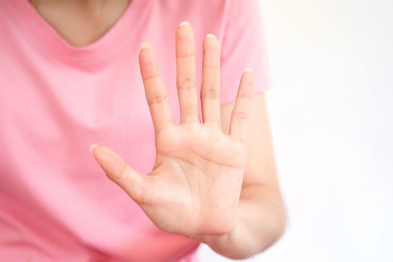 Mulher de camiseta rosa tem a mão aberta, em sinal de pare/basta com a violência de gênero. Ela tem a pele clara e não há imagem do rosto.