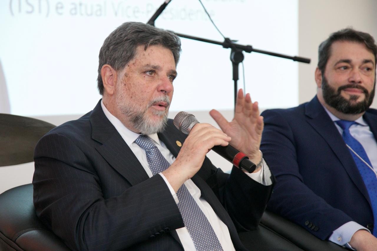 Ministro Caputo fala ao microfone gesticula. Ele é branco, de barba e cabelo grisalho, veste terno escuro com camisa branca e gravata cinza. A seu lado aparece o vice-presidente Alexandre Corrêa da Cruz.