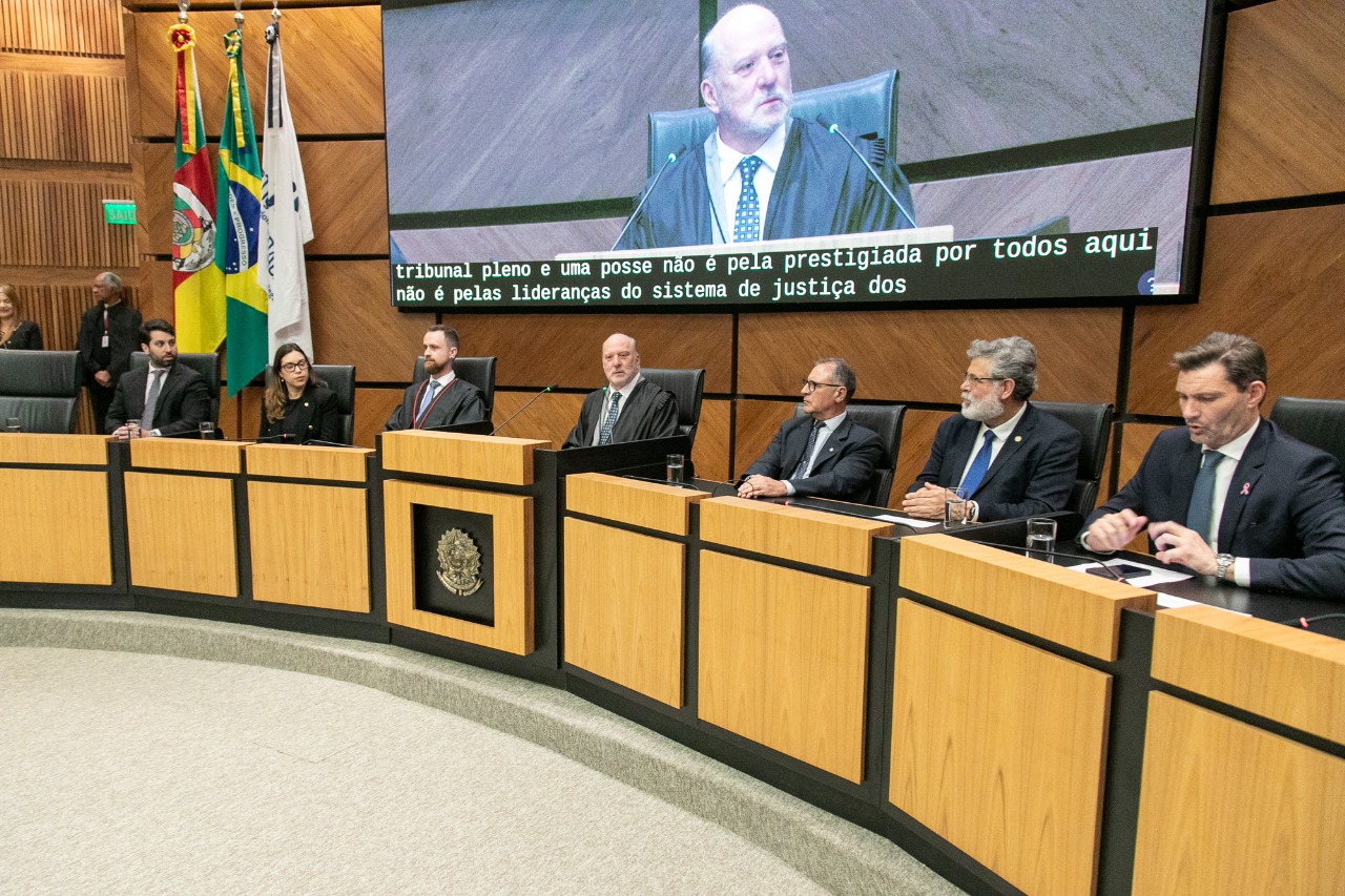 Sete pessoas aparecem na mesa oficial. Martins Costa está ao centro e também aparece falando no telão atrás da mesa. 