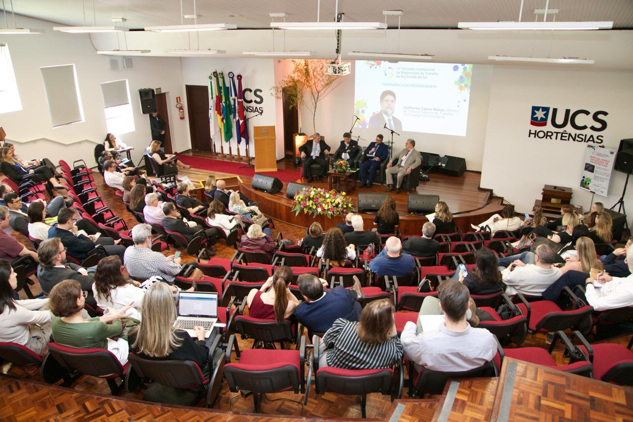 Vista aérea do auditório. Público aparece em primeiro plano, de costas. Ao fundo está o palco com quatro pessoas sentadas.