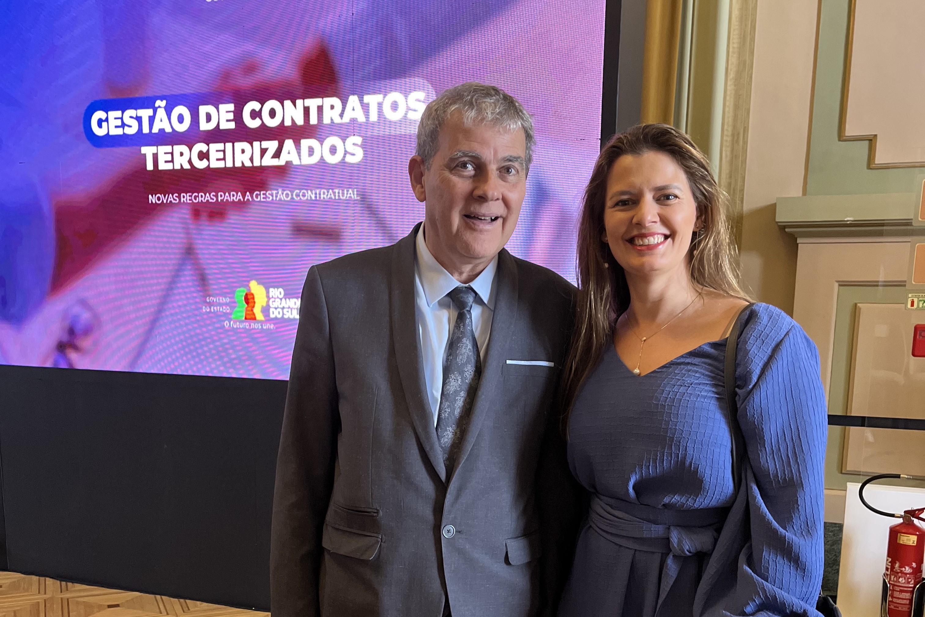 Ricardo e Andrea posam para a foto sorrindo. No lado esquerdo aparece um telão com o texto "Gestão de Contratos Terceirizados" sobre o logo do Governo do Estado do RS.