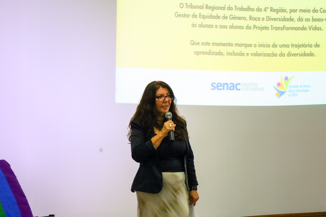 Juíza Lúcia fala ao microfone diante de uma tela de projeção. No fundo, o texto de boas-vindas do projeto “TransFormando Vidas”.