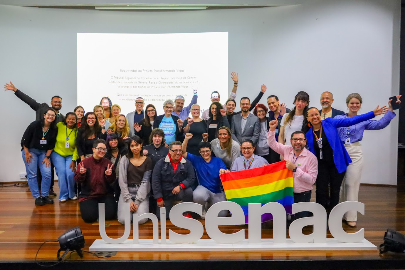 Grupo posa sorridente no auditório, com os braços erguidos em celebração. À frente, a palavra “UniSenac” em destaque e uma bandeira LGBT+.