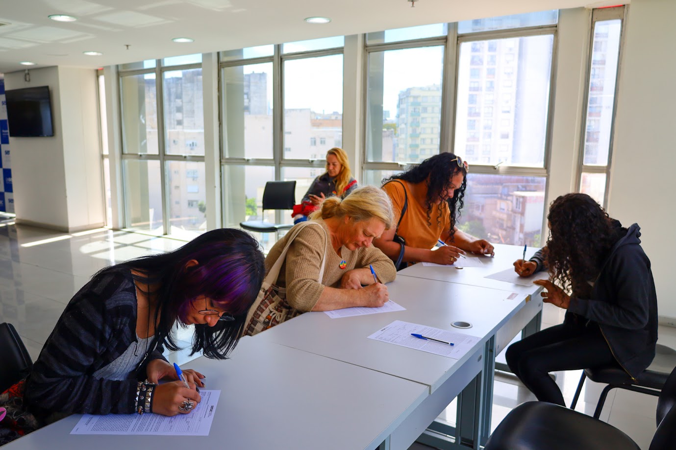 Estudantes do curso preenchendo os formulários. A cena se passa em uma sala iluminada por janelas amplas, com vista para prédios do centro de Porto Alegre. 