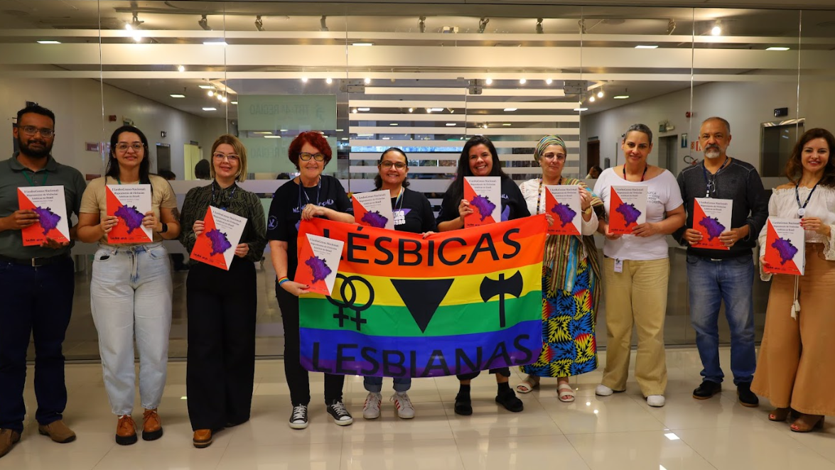 Participantes da reunião posam sorrindo, segurando os exemplares do LesboCenso Nacional. No centro, duas delas exibem uma bandeira colorida com os símbolos lésbicos e os dizeres “LÉSBICAS LESBIANAS”.