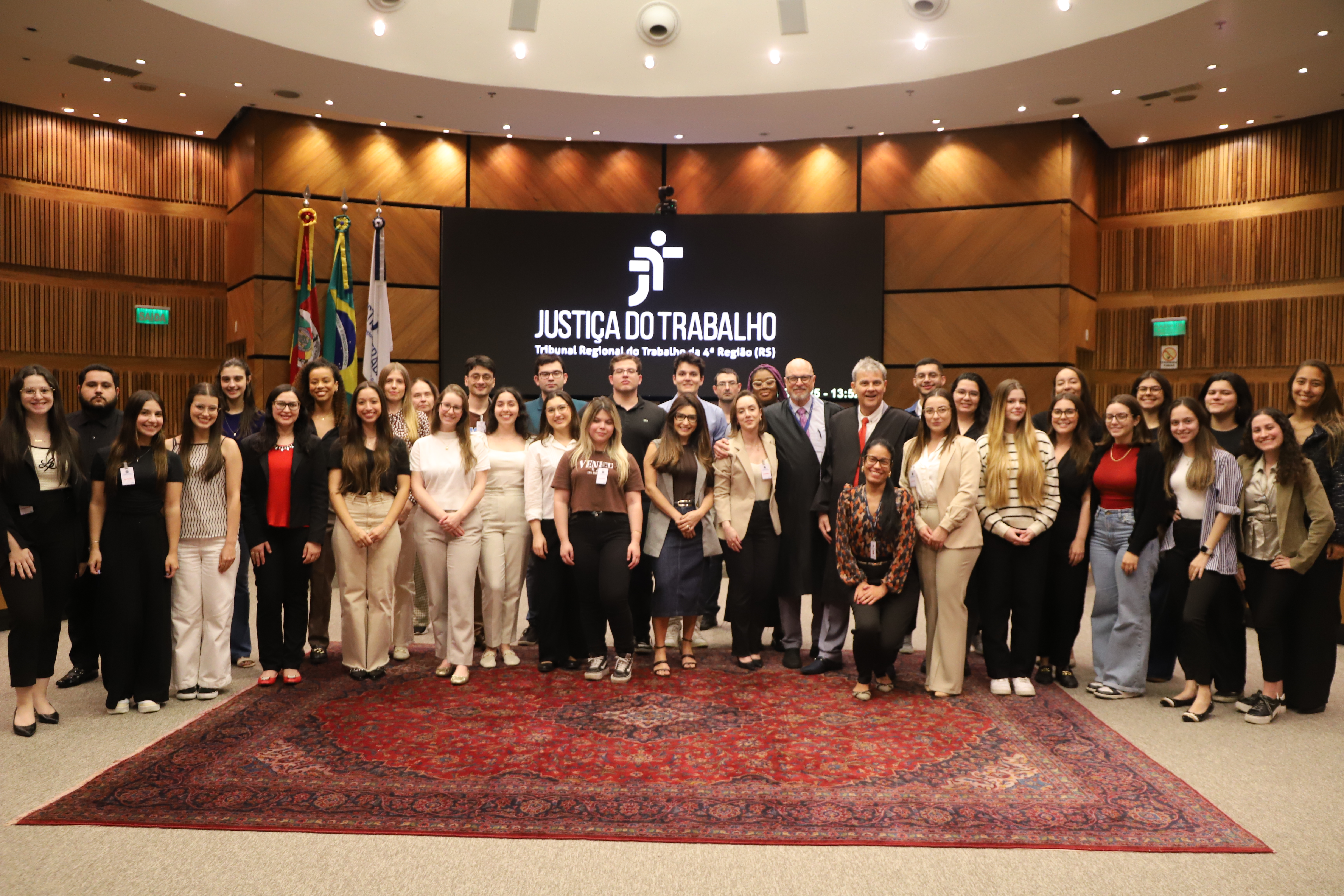 Grupo grande de pessoas posa para foto  no plenário do TRT-RS, com painel ao fundo exibindo “Justiça do Trabalho”.