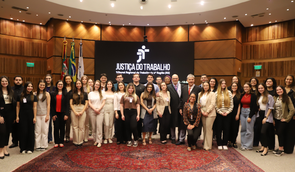 Grupo grande de pessoas posa para foto  no plenário do TRT-RS, com painel ao fundo exibindo “Justiça do Trabalho”.