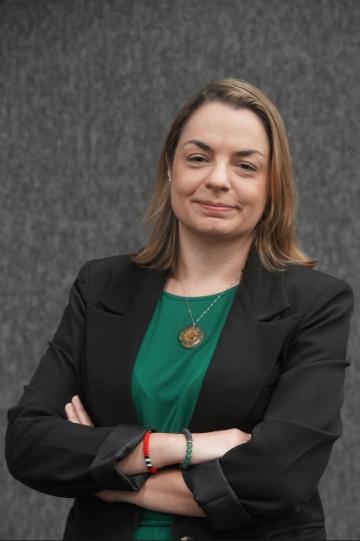 A juíza Gabriela é uma mulher de cerca de 45 anos. Tem e pele clara e cabelos castanhos e lisos, pela altura do ombro. Veste um blaser preto e blusa verde.