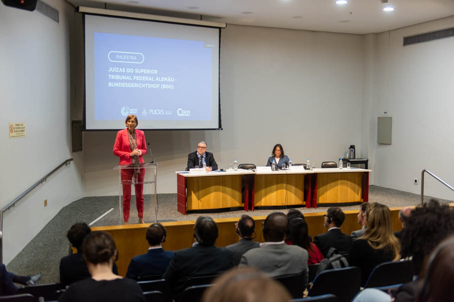 Palestra com mulher em terno vermelho no púlpito, tela sobre "Juízas do Tribunal Federal Alemão", painel com dois participantes e público na plateia.