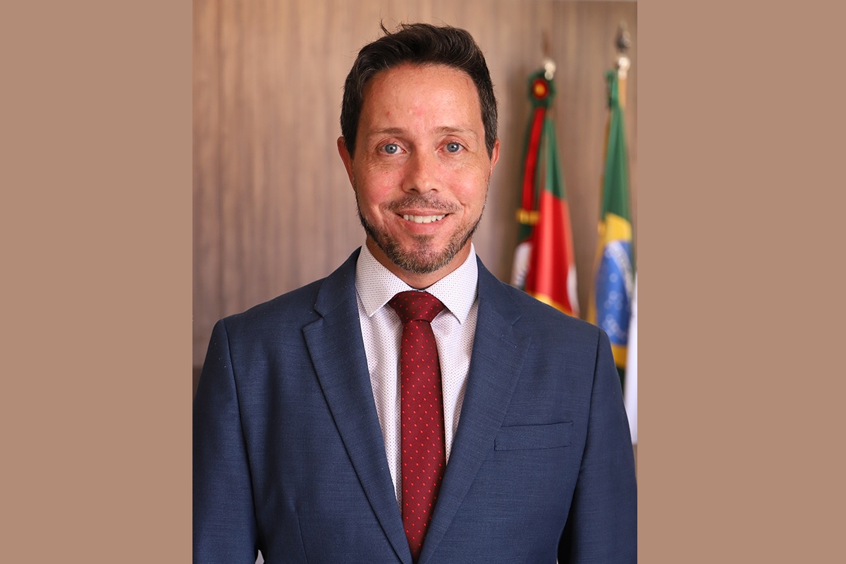 Rodrigo Trindade é branco, tem o cabelo liso escuro e curto e barba curta. Usa blazer azul marinho, camisa branca e gravata bordô. Está posando para a foto sorrindo. Atrás dele aparecem as bandeiras do RS e do Brasil.