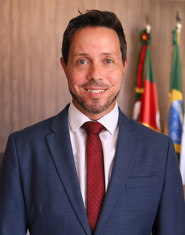 Rodrigo Trindade é branco, tem o cabelo liso escuro e curto e barba curta. Usa blazer azul marinho, camisa branca e gravata bordô. Está posando para a foto sorrindo. Atrás dele aparecem as bandeiras do RS e do Brasil.