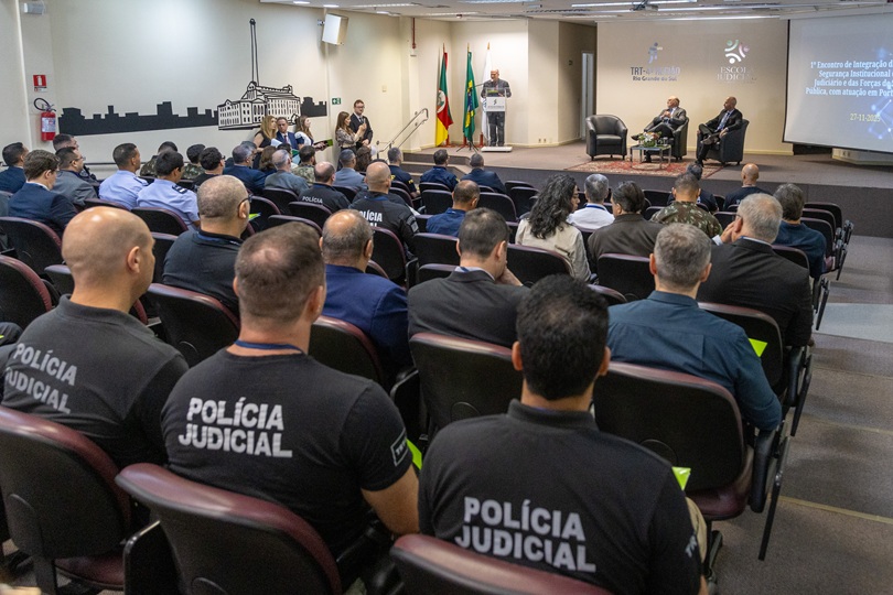 Foto em angulo aberto do auditório, mostrando público de costas e palco ao fundo. Em primeiro plano,três agentes aparecem com juma camiseta preta escrita "Polícia Judicial" nas costas.