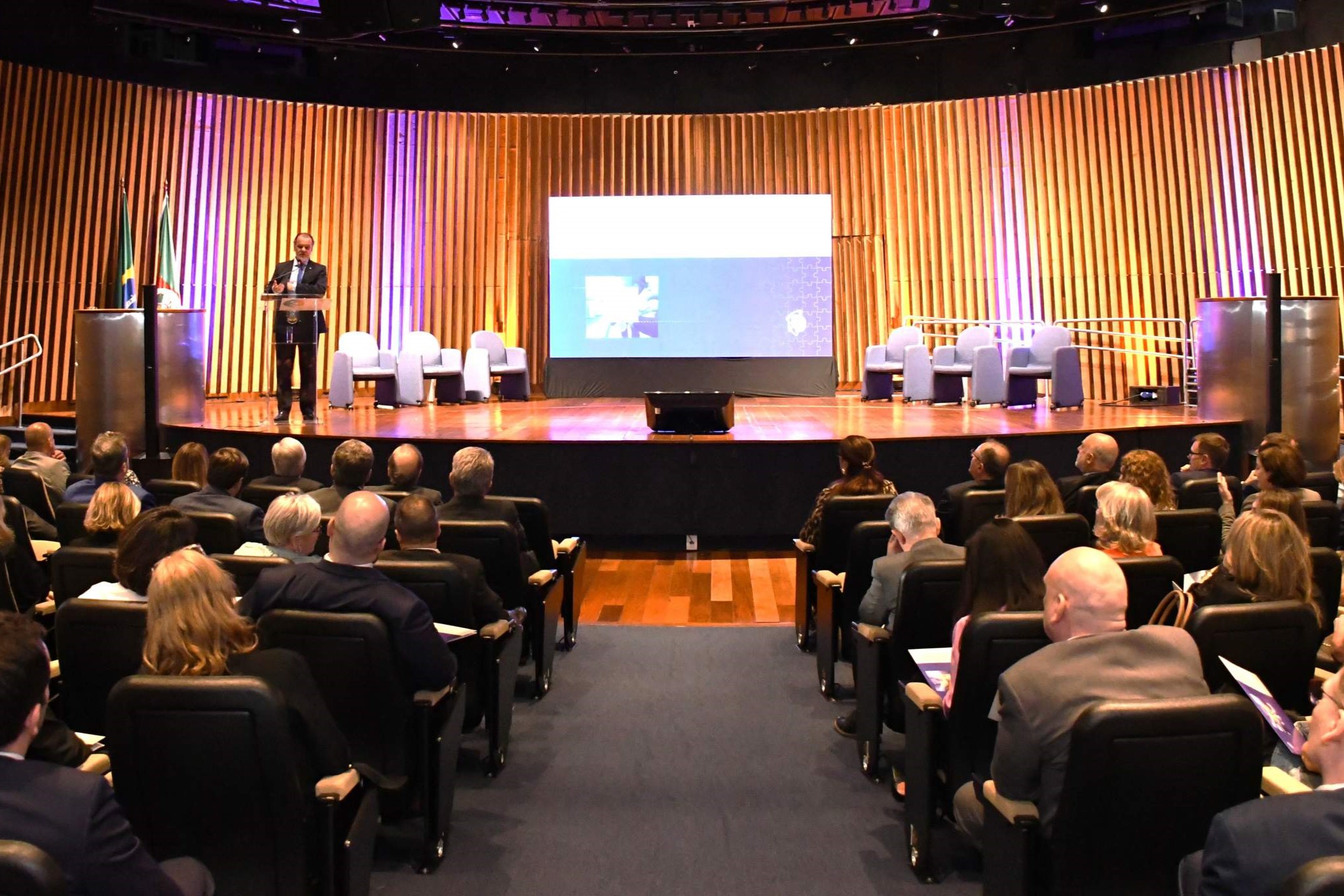 Foto geral do auditório do congresso. O público aparece de costas em primeiro plano, com um homem de pé no palco ao fundo.