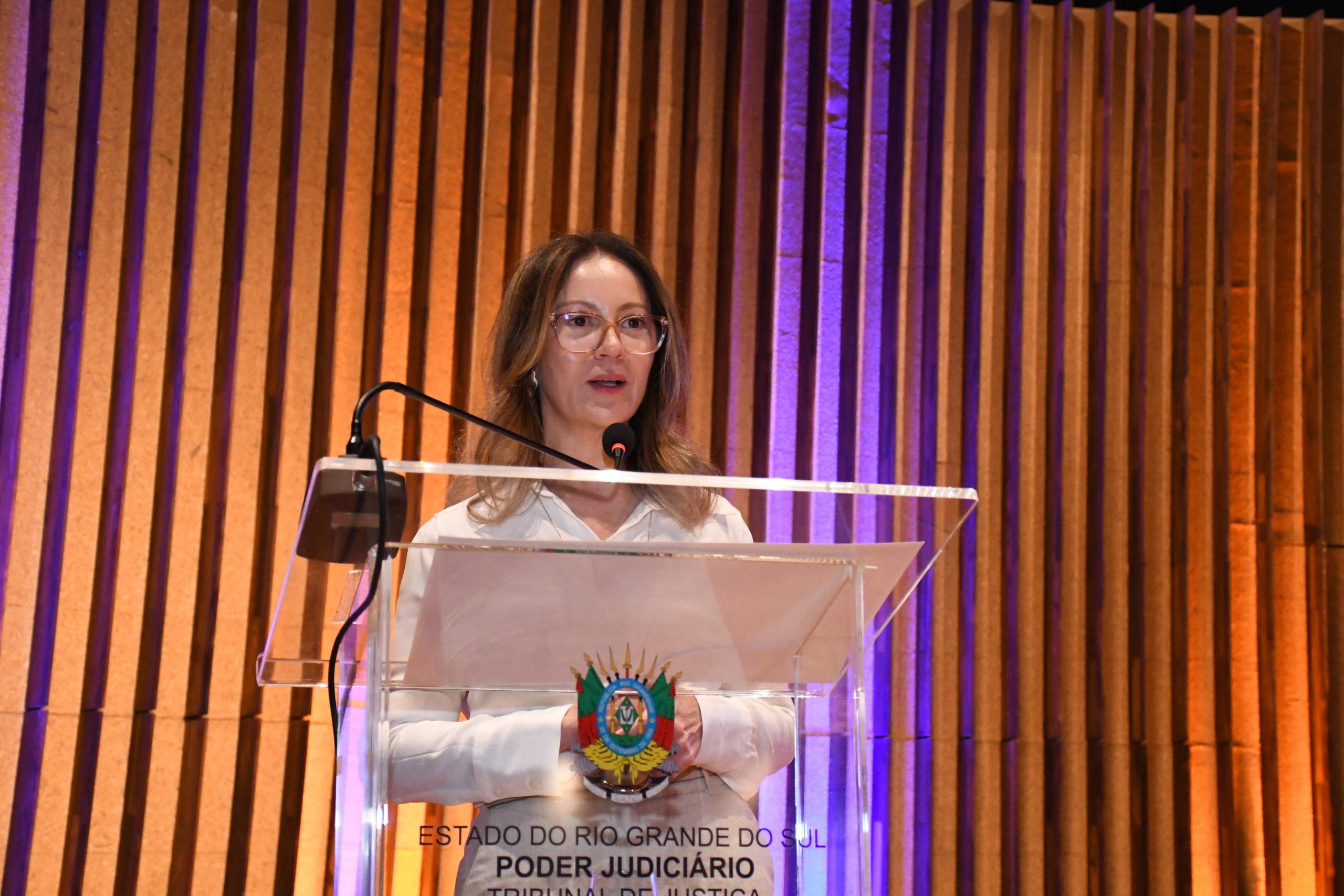 Adriana Fontoura é branca e de cabelos castanhos e lisos. Ela está de pé, falando atrás de uma tribuna. Na frete aparece o logo do Poder Judiciário do RS.