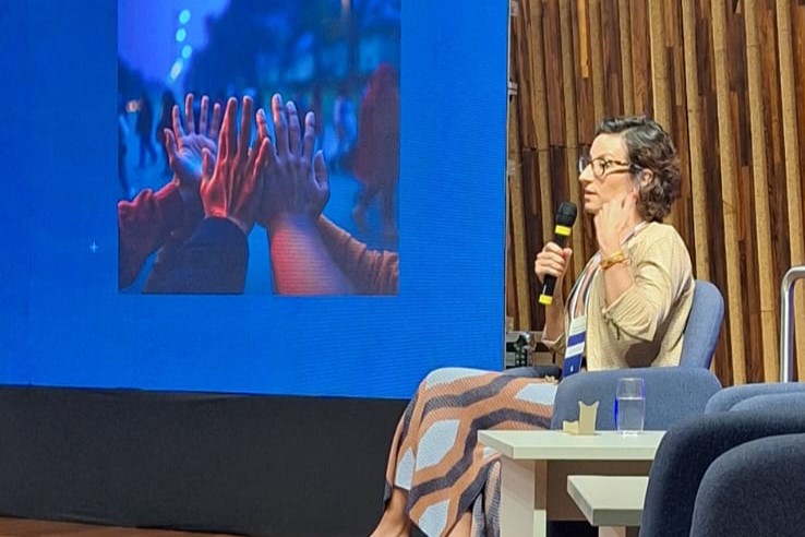 Mariana Lerina é branca, de cabelo castanho curto. Está sentada, falando e gesticulando de perfil. A seu lado aparece um telão com a imagem de várias mãos se tocando.