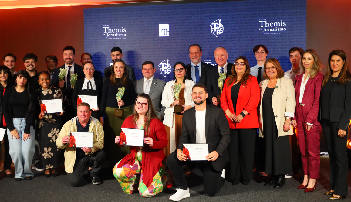 Grupo de premiados posa sorridente diante do painel “Prêmio Themis de Jornalismo – Poder Judiciário RS”. Alguns seguram troféus e certificados; todos estão em trajes sociais.