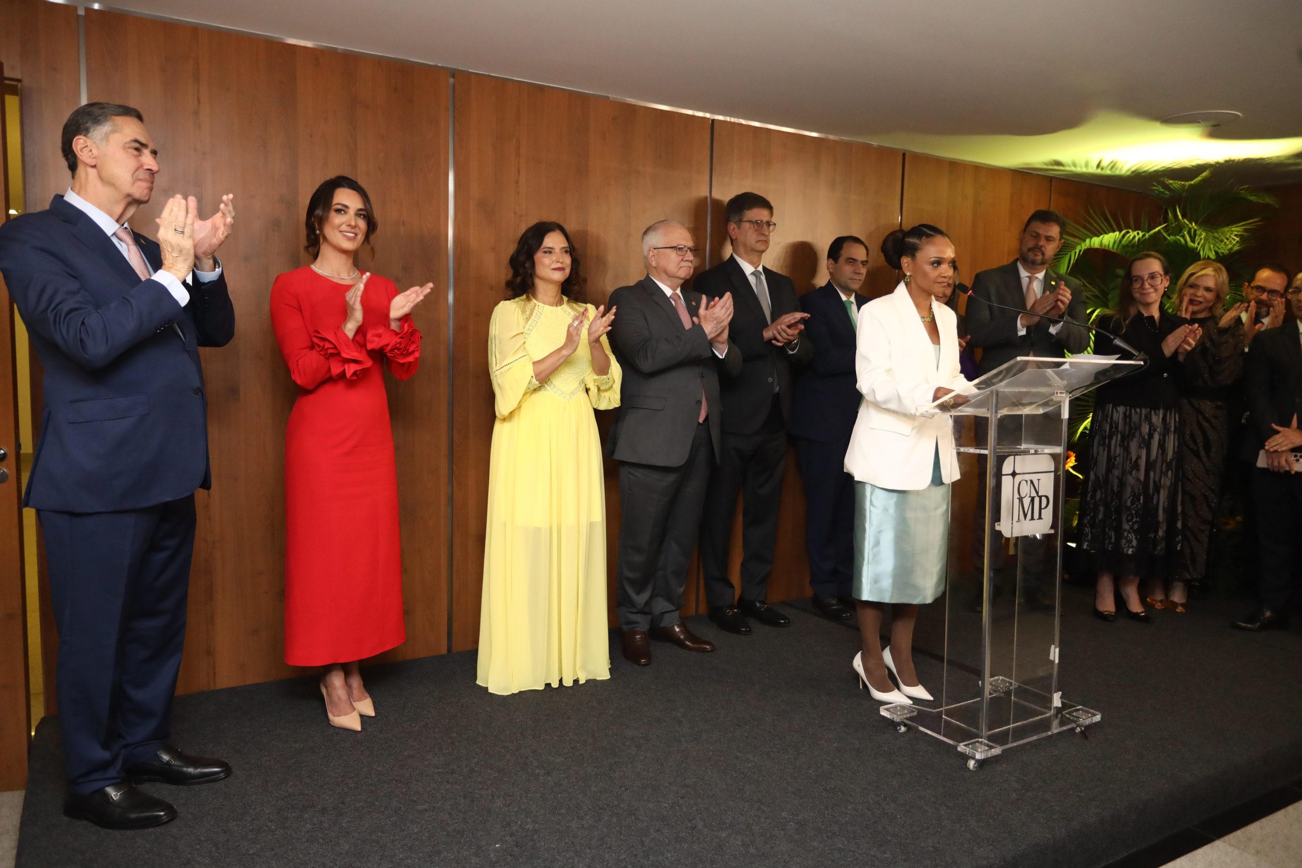 Juíza Karen Luise falando, de um púlpito. Atrás dela, autoridades perfiladas no dispositivo oficial, aplaudindo.