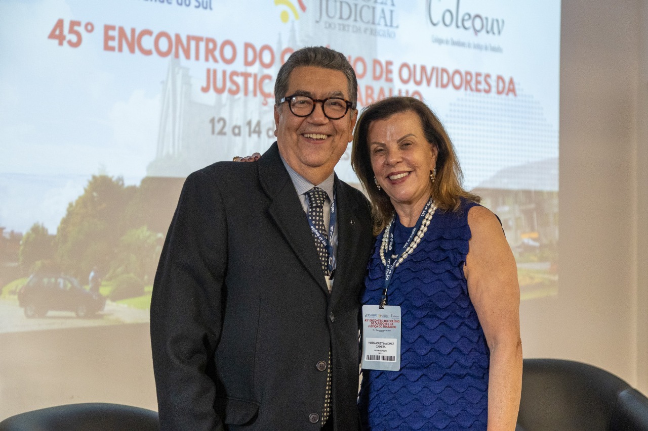 Jorge Ramos e Maria Caixeta posam para a foto sorrindo. Atrás deles aparece um telão com a arte do Encontro. 