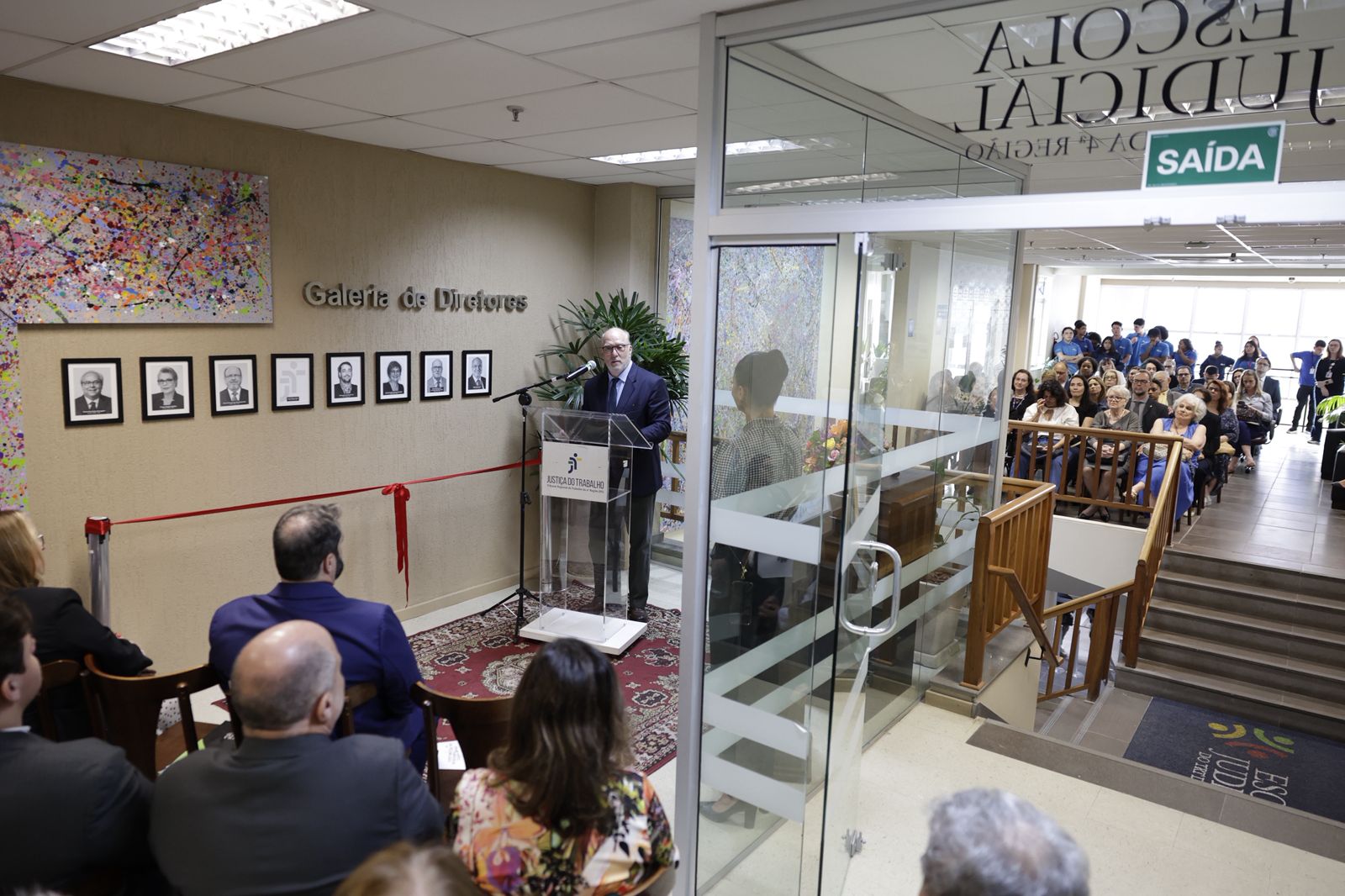 A foto mostra parede com fotos e uma pessoa discursando.