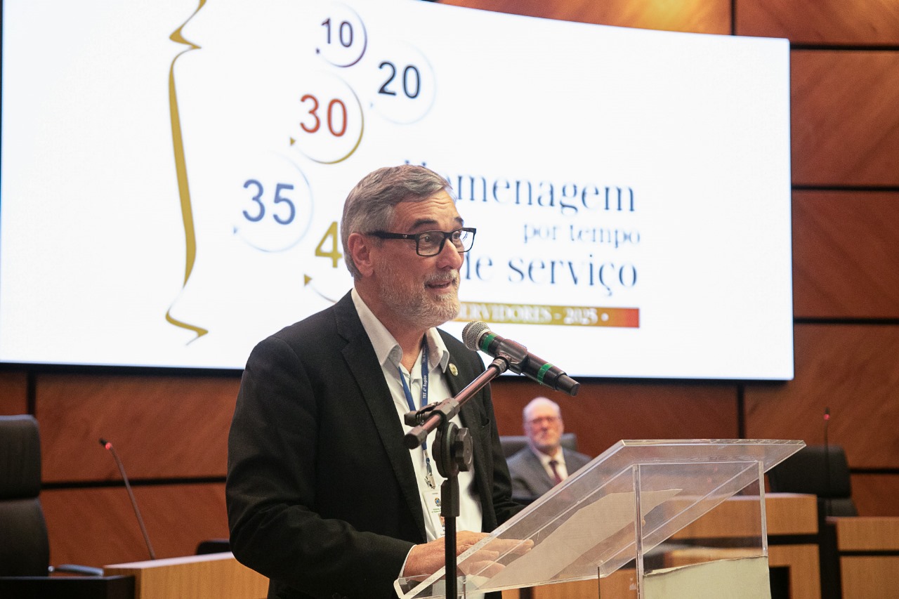Peixto é branco e grisalho, usa barba curta e veste terno. Fala ao microfone do púlpito de pé. atrás dele aparece o telão com a  arte do evento.